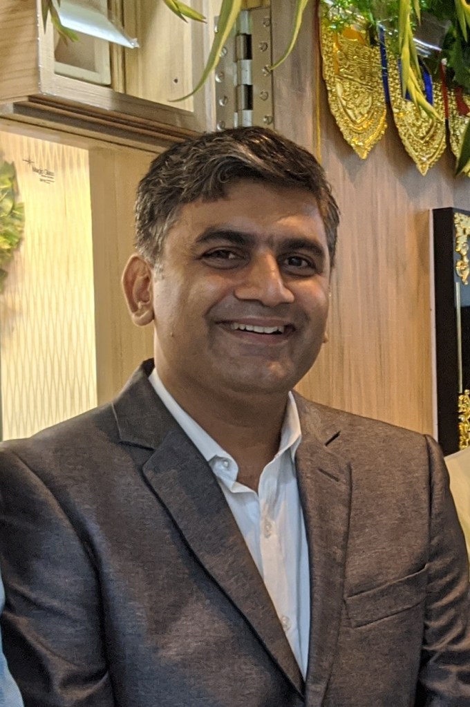 A smiling man wearing a grey suit and white shirt, standing in a well-decorated indoor setting with plants in the background.
